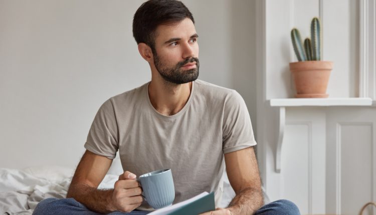 Indoor shot of pensive young Caucasian guy with thick beard, being deep in thoughts after reading life story, holds book and cup of tea, poses on bed against domestic interior. Leisure concept.